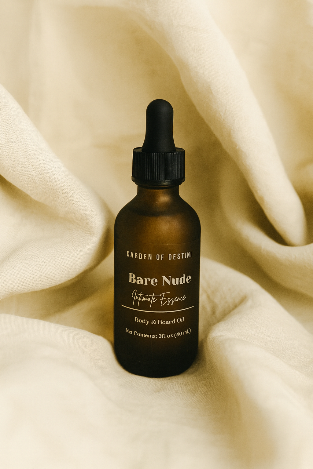A 2oz amber glass dropper bottle of Garden of Destini Bare Nude Intimate Essence Body & Beard Oil rests on soft beige fabric. The folds of the fabric create a warm, elegant backdrop that highlights the minimalist white label and luxurious aesthetic of the product.